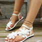 White Zephyra flat sandals adorned with delicate layered floral lace appliques on straps, worn on feet with white toenails and gold anklets, set against a rustic wooden background.