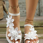 White floral lace flat sandals worn on feet with white toenails, featuring delicate appliques and an ankle strap, set against a rustic wooden background.