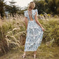 Woman seen from the back wearing a white Zephyrin bohemian midi dress with blue floral print, featuring ruffled cap sleeves and an open back tied with a ribbon, set in a natural outdoor scene.