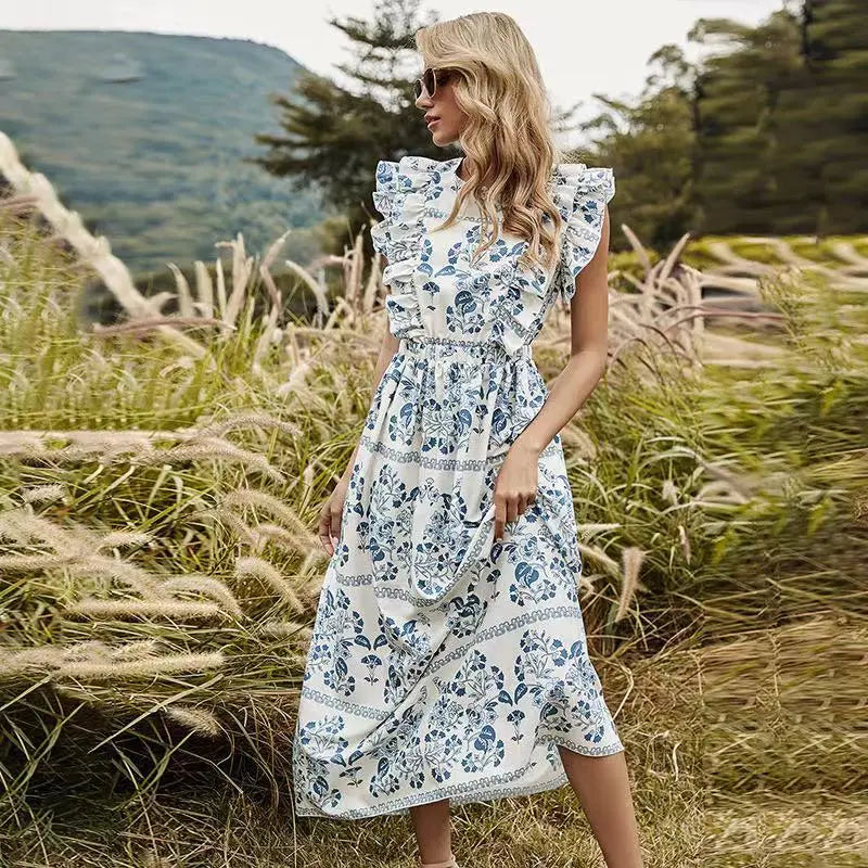 A woman models a white midi dress with a blue floral print, ruffled cap sleeves, and an elasticated waist, standing in a grassy, natural outdoor landscape.