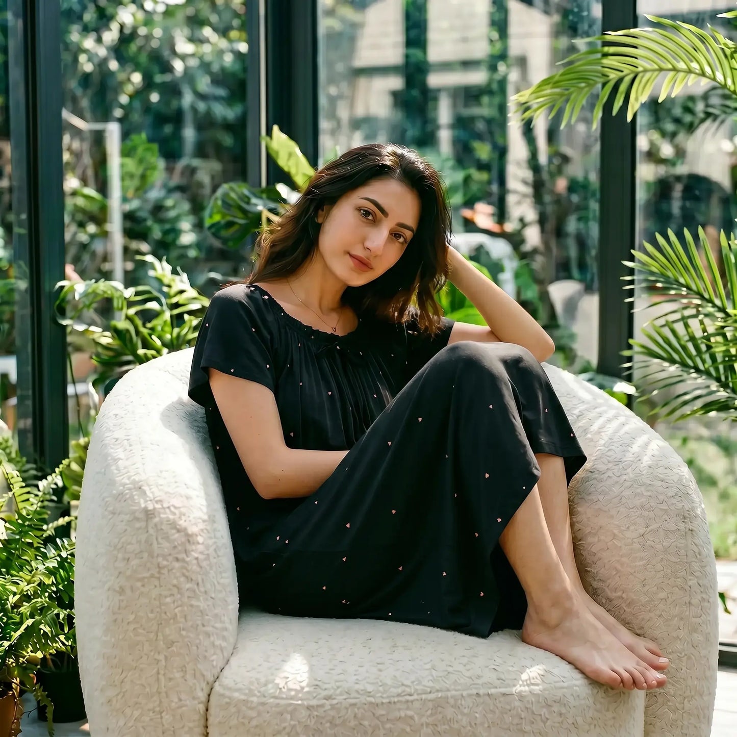 Model wears Zevryn black modal lounge dress, with gathered scoop neck, flutter sleeves, and pink micro-pointelle hearts, relaxing in a sunny conservatory.