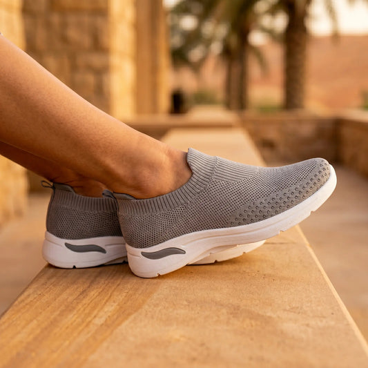 Elegant grey knit slip-on walking sneakers with a breathable upper and thick white sole, worn by a woman relaxing outdoors on a stone ledge.