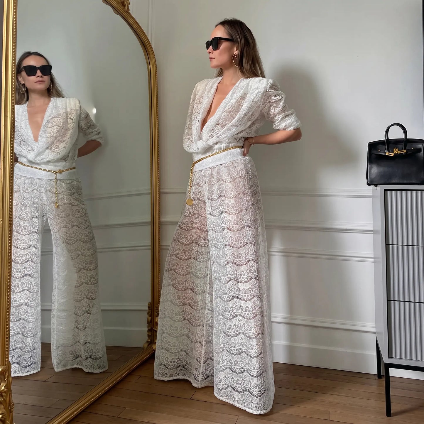 A woman models a Zhyra white lace two-piece set featuring a draped cowl neck top with puff sleeves and flowing wide-leg pants, styled with a gold chain belt.
