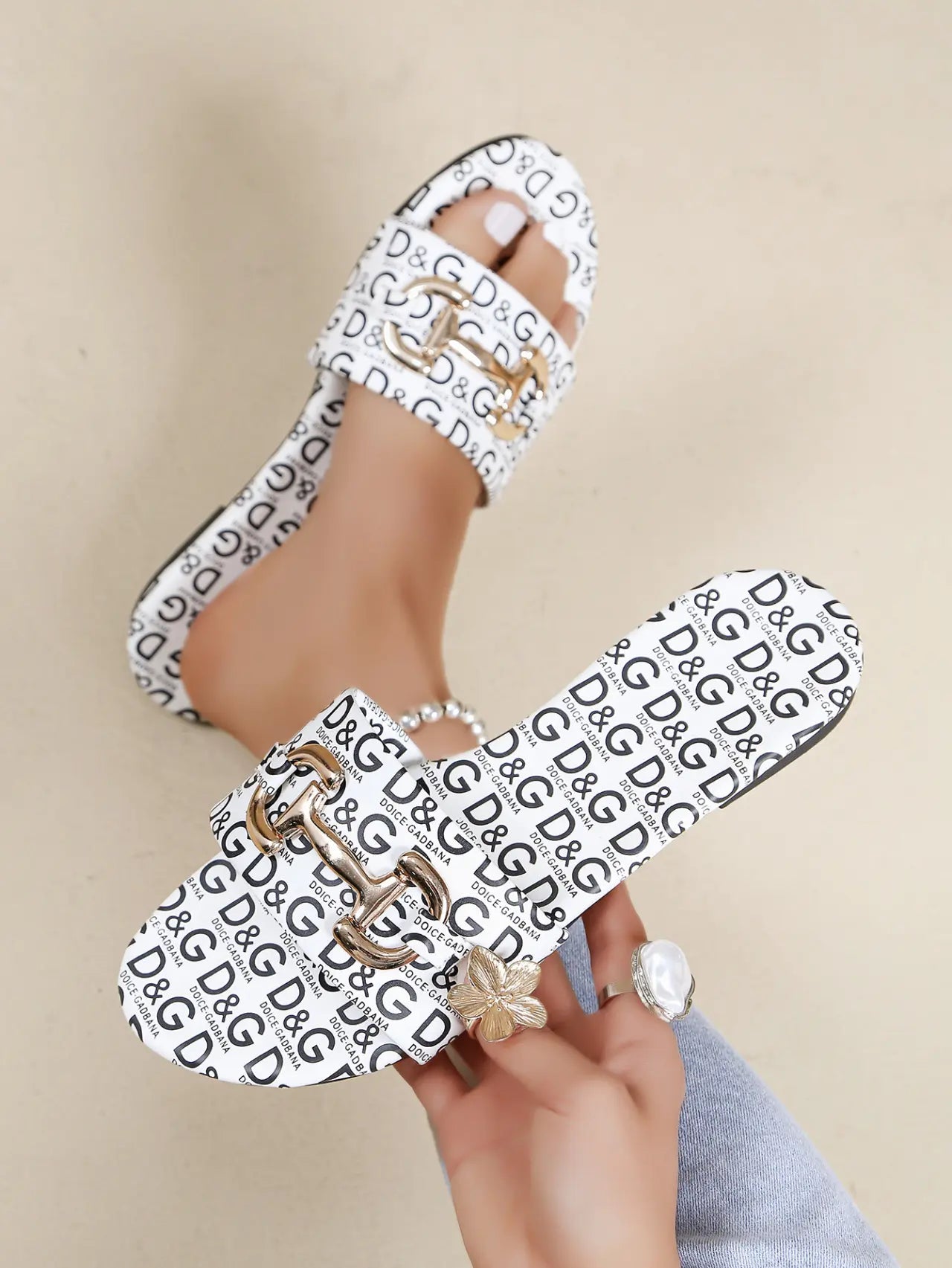 Zirali women's white slide sandals featuring an all-over black logo print and ornate gold-tone buckle hardware, shown on a foot with rings and held by a hand.