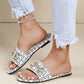 A woman's feet wearing white Zirali logo slide sandals with a black monogram print and ornate gold-tone buckles, paired with light-wash distressed denim jeans.