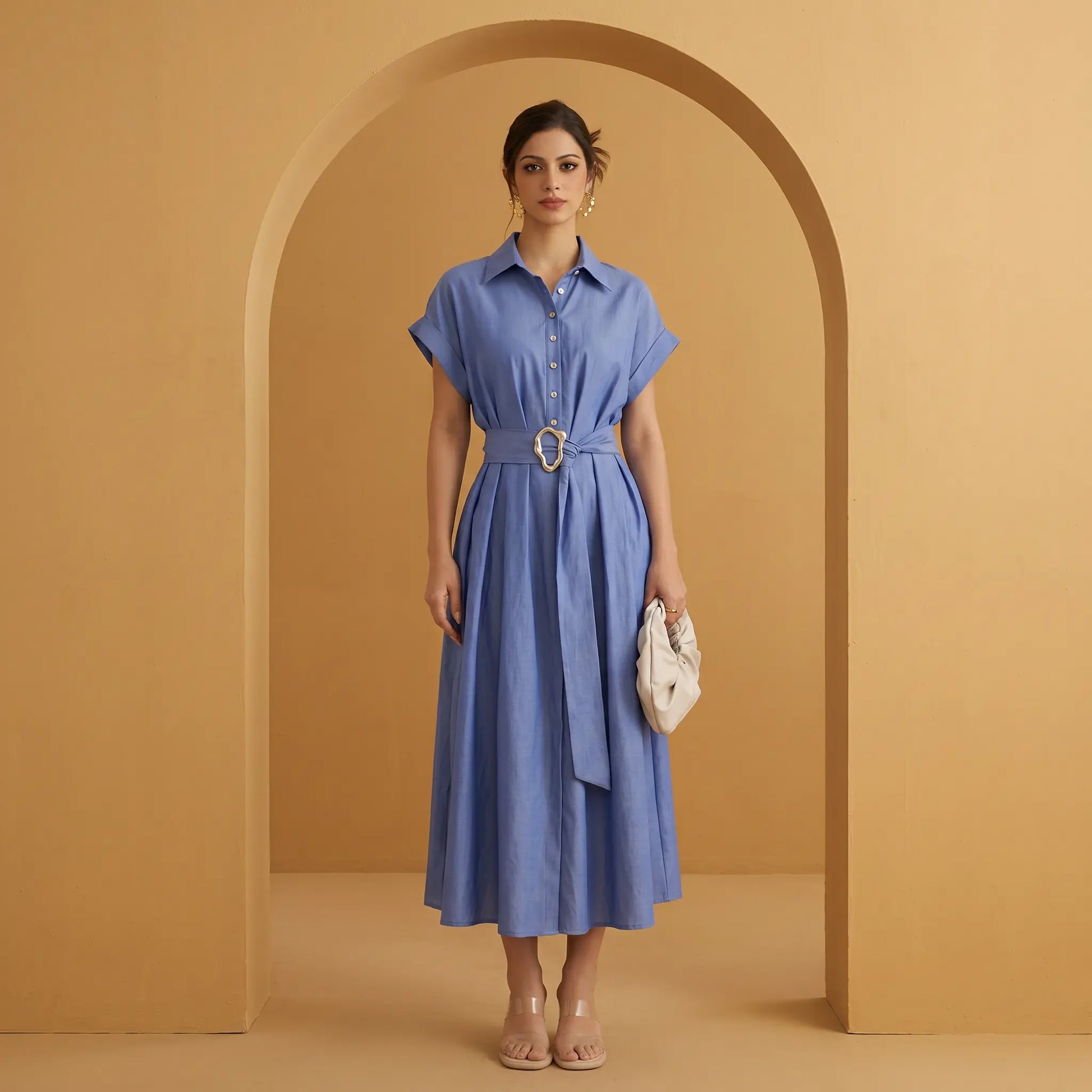 Model wearing a Zivro chambray blue linen-blend A-line midi dress, featuring a point collar, cuffed short sleeves, and a wide fabric belt with an organic gold-tone buckle, presented against a warm studio archway.
