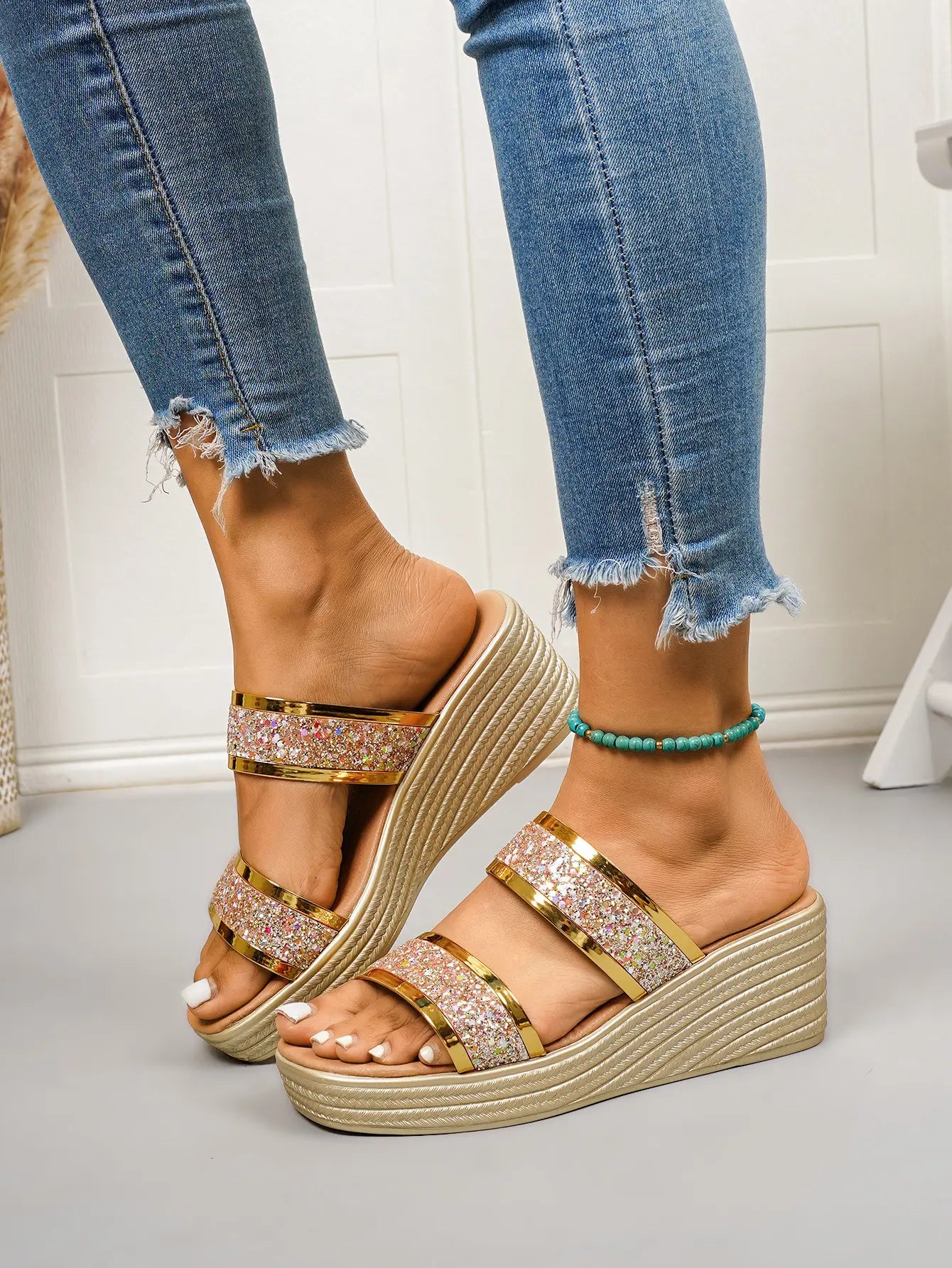 Zlata Women's gold glitter wedge slide sandals worn by a model with distressed denim jeans, showcasing the sparkling rose gold and iridescent straps and textured wedge heel.