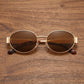 Gold metal oval frame sunglasses with dark amber brown tinted lenses and tortoise shell temple tips, displayed on a wooden surface.