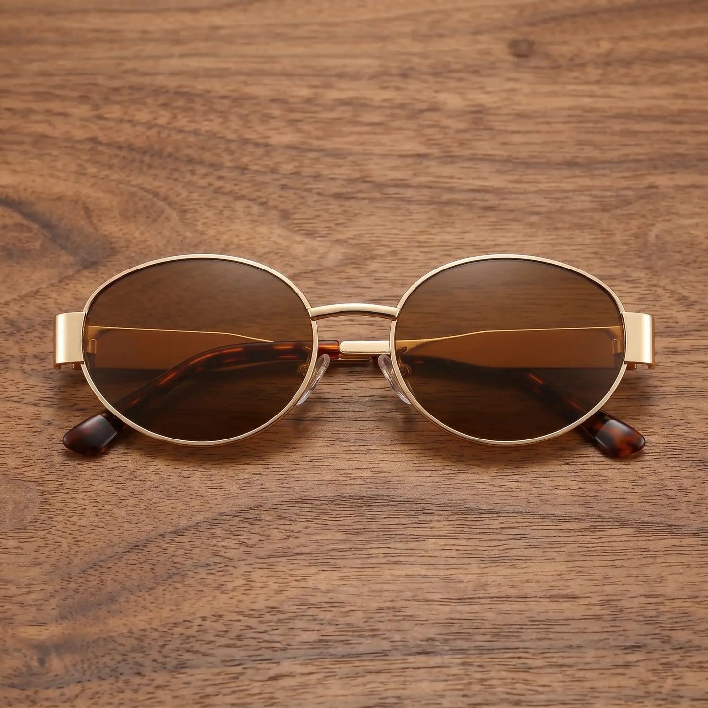 Gold metal oval frame sunglasses with dark amber brown tinted lenses and tortoise shell temple tips, displayed on a wooden surface.