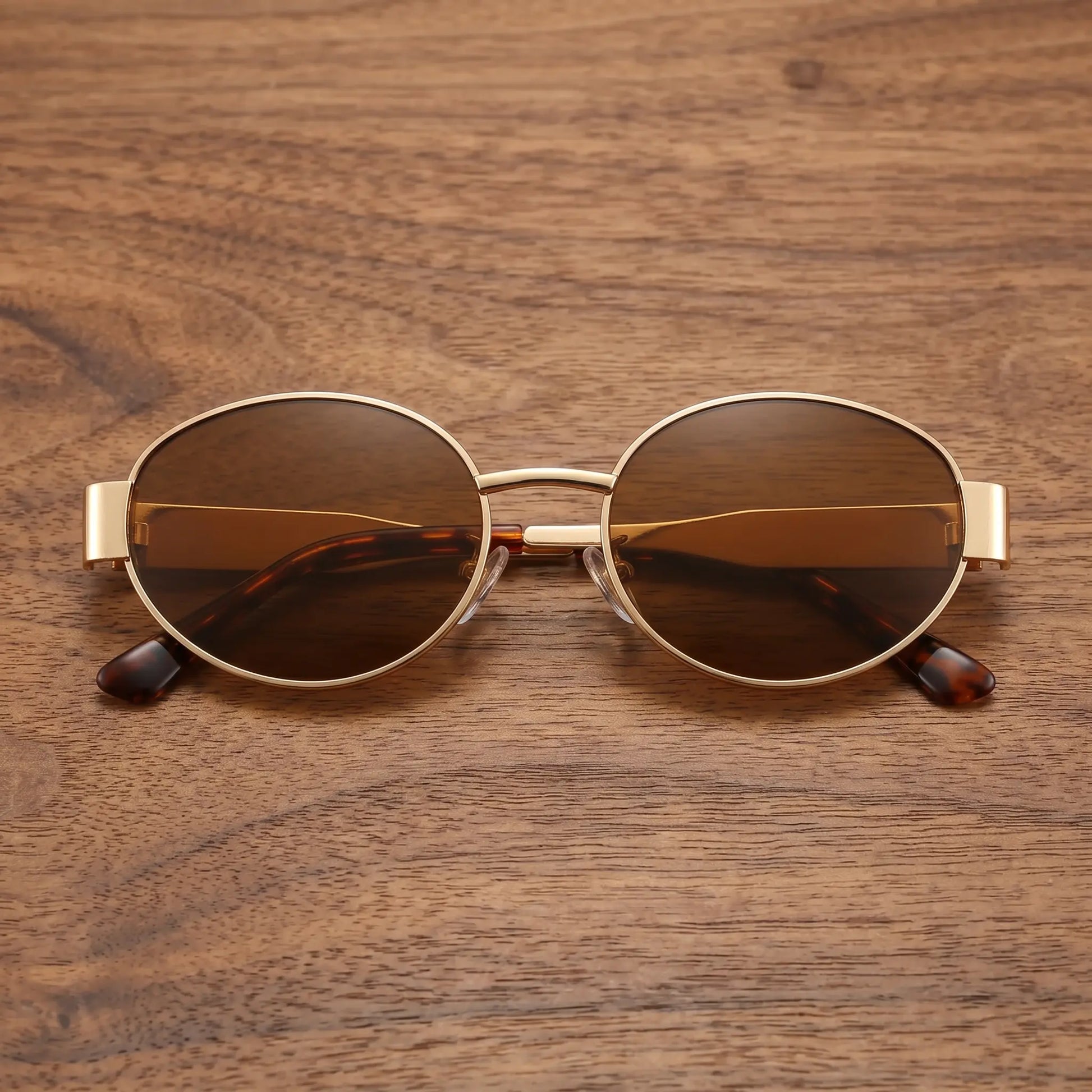 Gold metal oval frame sunglasses with dark amber brown tinted lenses and tortoise shell temple tips, displayed on a wooden surface.