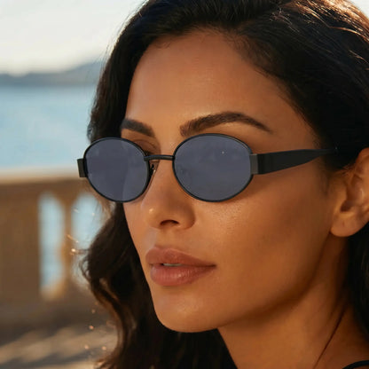 Close-up profile of a woman wearing Zokari Obsidian Black Oval Metal Frame Sunglasses with dark grey lenses, set against a blurred outdoor coastal background.