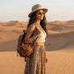 Model wearing Zokari gold oval metal frame sunglasses, straw hat, and bohemian ensemble with a leather backpack in a serene desert setting.