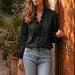 Woman models a Zokiru Midnight Black Ruffled V-Neck Blouse with long blouson sleeves, styled with light wash jeans, standing outdoors by an ornate wooden door and potted plants. The elegant cascading ruffles frame the neckline.