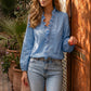 Woman wears a sky blue Zokiru Ruffled V-Neck Blouse with cascading ruffles and long sleeves, paired with jeans in a romantic outdoor setting.