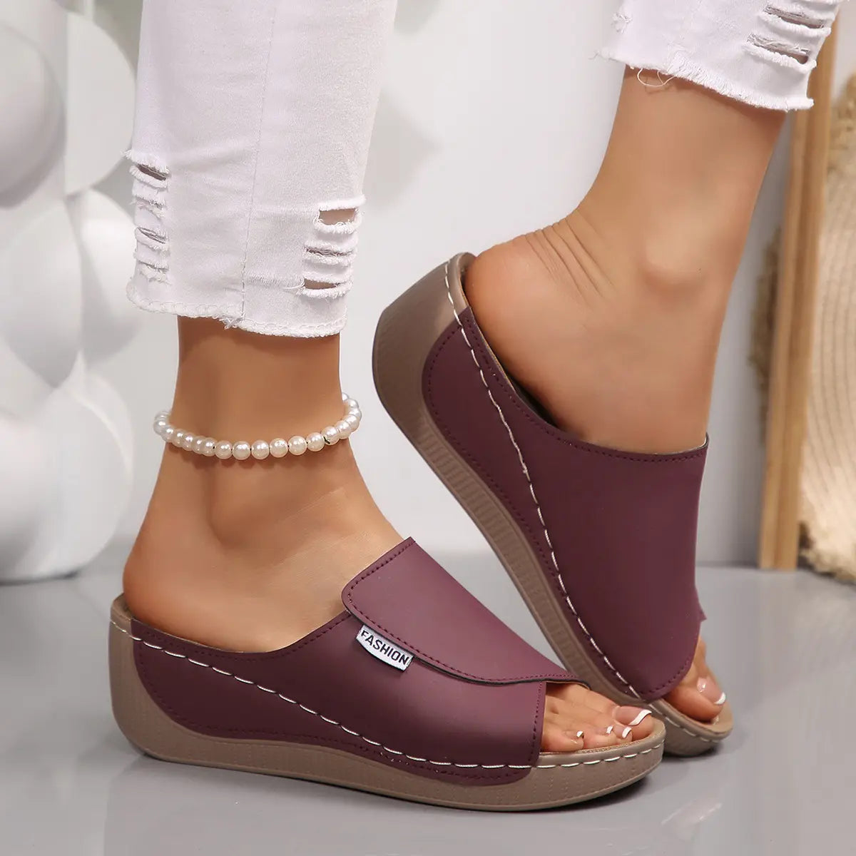 Women's wine red slide platform sandals with an open toe, single wide strap, and layered wedge sole, styled with ripped white jeans and a pearl anklet. Features contrast stitching and a small 'FASHION' label.