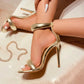 Gold Zoltra elegant stiletto sandals with puffy straps on a woman's feet, showing a side view with a decorative chain accent.
