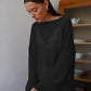 Female model wearing a Zomari black semi-sheer knit sweater with long bell sleeves and a wide boat neck in a relaxed indoor setting.