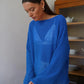 Model wearing a vibrant blue Zomari sheer bell sleeve knit sweater with an open-knit texture and boat neck, styled in a relaxed indoor setting.