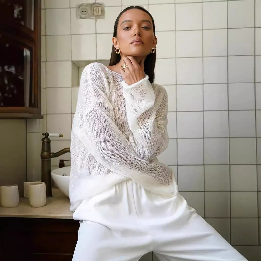 Female model poses wearing a Zomari Pristine White sheer knit sweater, showcasing its relaxed fit, wide boat neckline, and translucent open-knit texture. This lifestyle image highlights its effortless style.