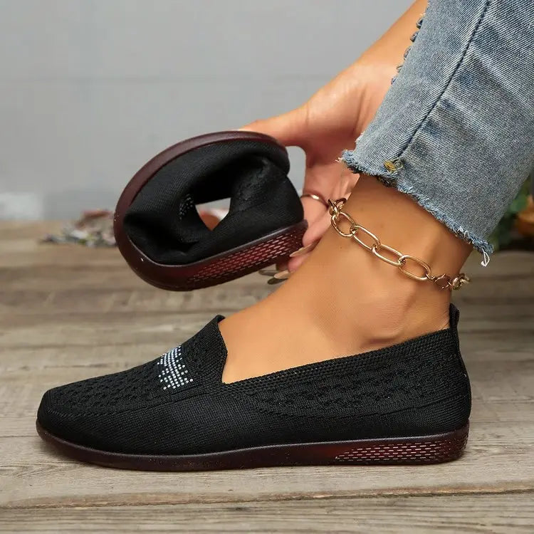Black Zoria knit loafers with crystal embellishments on the vamp, shown on a woman's foot with denim jeans against a wooden floor.