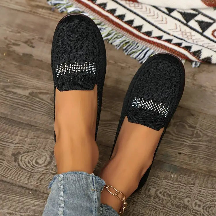 A pair of black Zoria breathable knit loafers with crystal embellishments on the vamp, worn on feet with distressed denim jeans and a gold anklet, styled on a wooden floor.