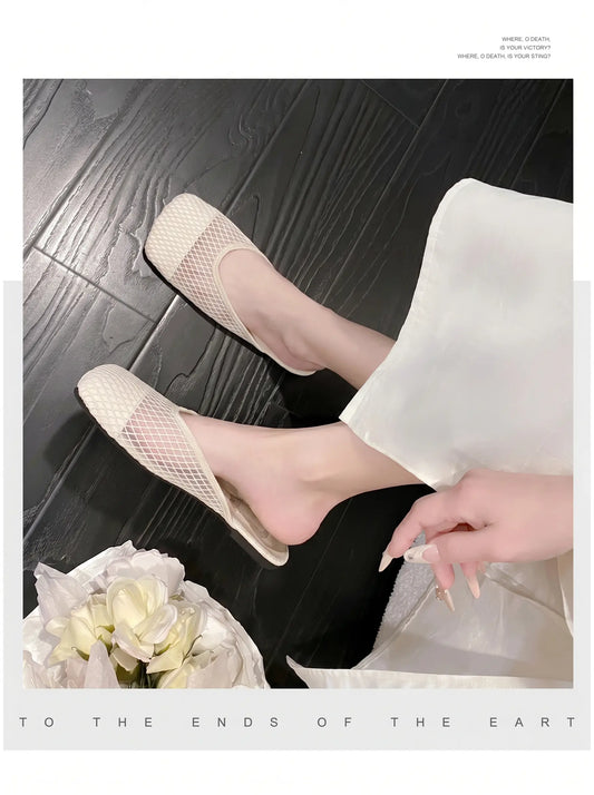 A pair of soft off-white mesh mules with a square toe, worn on a foot against a dark wood floor and styled with white fabric and flowers. Features airy diamond-patterned mesh and a subtle insole detail.