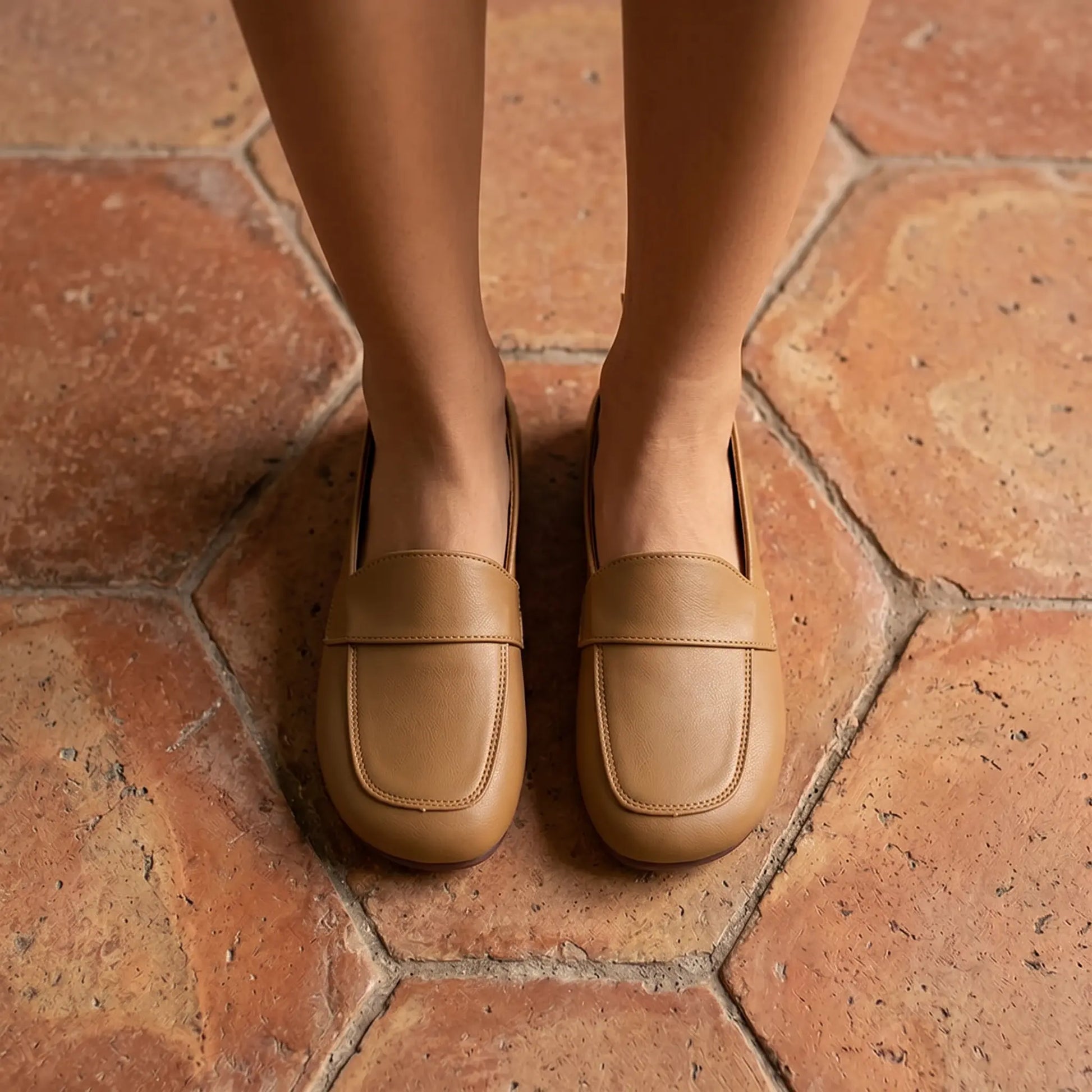 Elegant Zoryn 'Soft Apricot' vegan leather slip-on loafers, featuring a classic transverse vamp strap and round toe, worn by a woman standing on textured terracotta tiles.