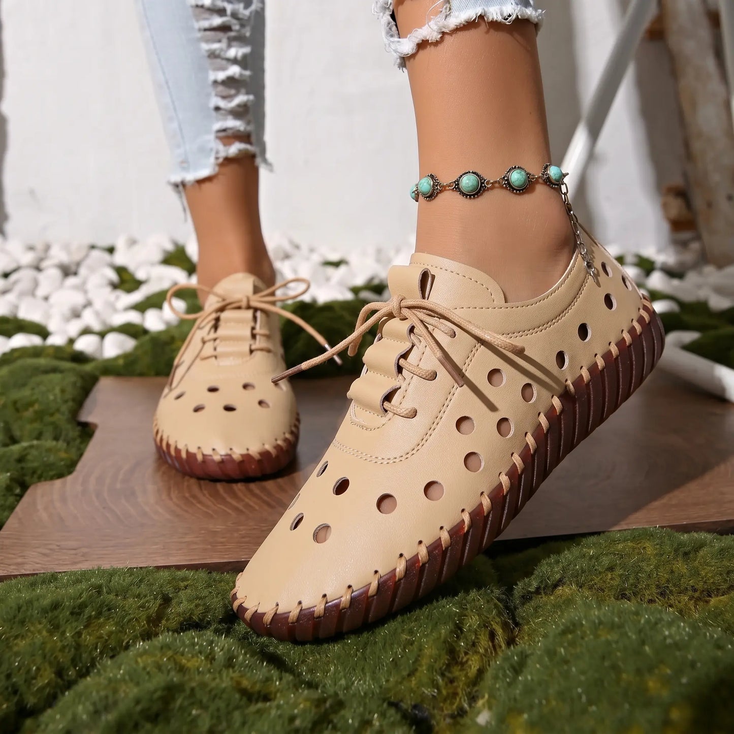 Lifestyle shot of Zoryn women's soft beige perforated lace-up flat loafers on a foot wearing distressed jeans and a turquoise anklet, styled with natural elements.