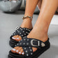 Black Zoryn platform slide sandals adorned with all-over silver studs and an ornate silver buckle, worn on a model's feet with white nail polish and a gold chain bracelet.