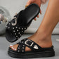 Zoryn black denim-look platform slide sandals adorned with silver studs and an ornate silver buckle, shown on a model's foot with a disco ball in the background.