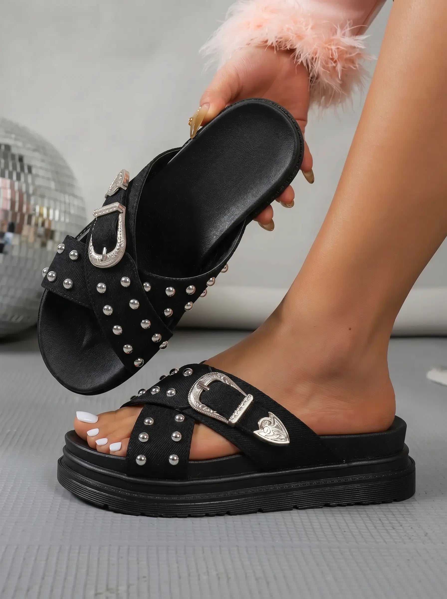Zoryn black denim-look platform slide sandals adorned with silver studs and an ornate silver buckle, shown on a model's foot with a disco ball in the background.