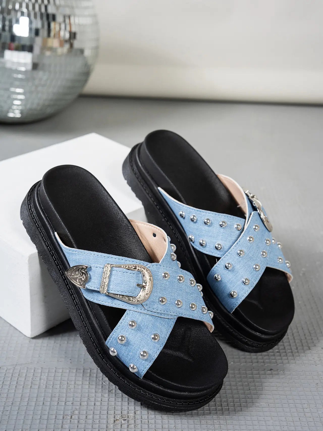 Zoryn studded platform slide sandals in light blue denim-look textile with ornate silver buckles and thick black soles. Displayed at a three-quarter angle on a studio floor.