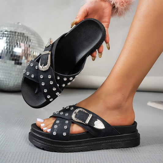 Zoryn black platform slide sandals adorned with silver studs and an ornate silver buckle, shown on a model's foot in a studio setting. Features crossed straps and a thick platform sole.