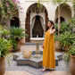 Model wearing a Zovith golden-yellow tent maxi dress with vertical stripes, spaghetti straps, and cream tassel hem, in a sunny Moroccan Riad.