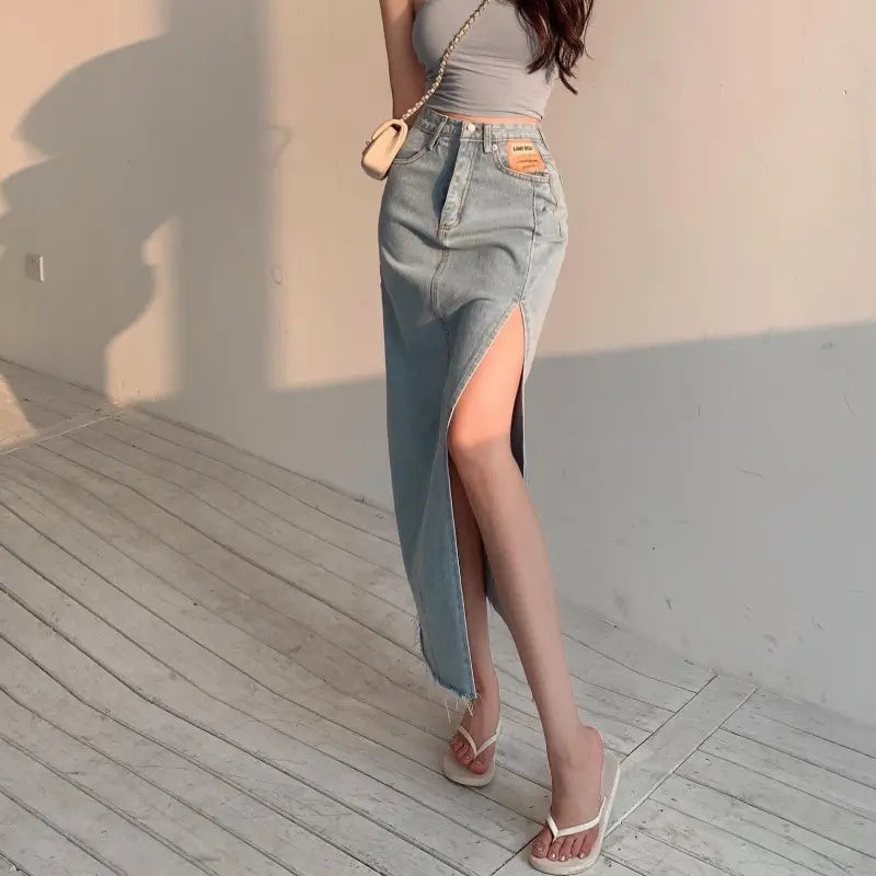 Model wearing Zubari vintage light blue high-waisted denim maxi skirt with dramatic thigh slit, raw frayed hem, and tan pocket patch.