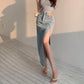 Model wearing a Zubari vintage light blue high-waisted denim maxi skirt. This lifestyle image highlights the dramatic side slit, raw frayed hem, and distinctive tan front pocket patch, showcasing the elegant A-line silhouette.