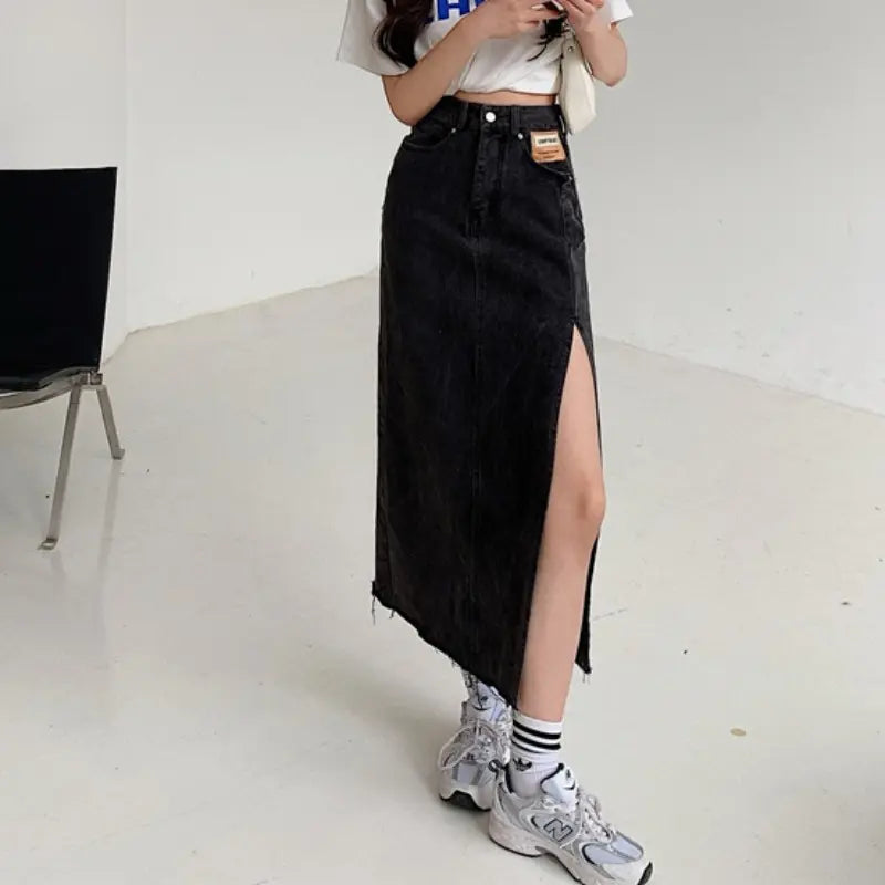 Model wearing a Zubari high-waisted A-line washed obsidian denim maxi skirt with a dramatic off-center thigh-high slit and a raw frayed hem. The skirt features a distinct tan patch on the left front pocket. Styled with a white crop top and sneakers.