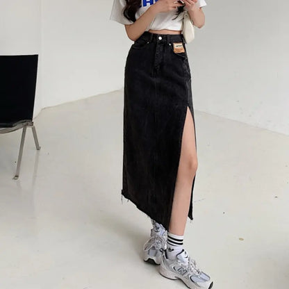 Model wearing a Zubari high-waisted A-line washed obsidian denim maxi skirt with a dramatic off-center thigh-high slit and a raw frayed hem. The skirt features a distinct tan patch on the left front pocket. Styled with a white crop top and sneakers.