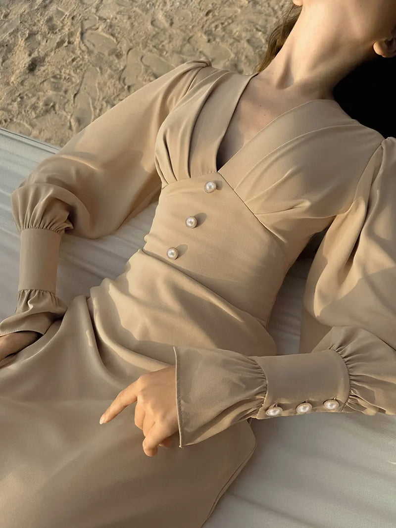 Elegant beige chiffon midi dress featuring a romantic V-neckline, voluminous bishop sleeves with pearl button cuffs, and a fitted waist. Shown on a model resting against a textured sandy background.