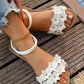 Woman's feet wearing white Zydra bohemian floral flat sandals with rhinestone accents, paired with distressed jeans and a gold anklet, resting on a wooden deck near water.