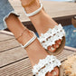 Close-up of white Zydra bohemian floral flat sandals worn by a woman, featuring delicate fabric flowers, sparkling rhinestones, and an adjustable ankle strap, shown on a wooden deck near water.
