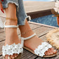 White Zydra bohemian flat sandals adorned with layered fabric flowers and small rhinestones, worn with ankle bracelets on a wooden deck by a pool. Featuring an adjustable ankle strap for a chic summer look.