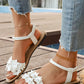 Women's white bohemian floral flat sandals with delicate fabric flower embellishments and a secure ankle strap, shown in a lifestyle setting on a wooden deck near water.