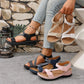 A pair of black comfort sport sandals with criss-cross elastic straps is worn on a model's foot, styled with light-wash denim jeans. Several other pairs in different colors are displayed on a concrete floor, with a stone wall background.