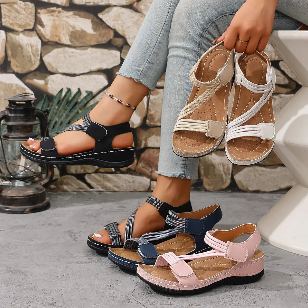 A pair of black comfort sport sandals with criss-cross elastic straps is worn on a model's foot, styled with light-wash denim jeans. Several other pairs in different colors are displayed on a concrete floor, with a stone wall background.