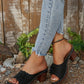 Black Zylaya wedge sandals with layered floral applique and perforated straps, shown on a person wearing jeans with a rustic wooden background.