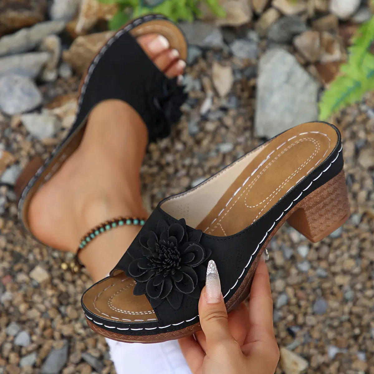Black Zylaya wedge sandals for women, featuring a decorative floral applique and perforated straps, shown on a foot with a natural pebble and stone background.
