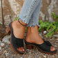 Black Zylaya women's wedge sandals with intricate floral embellishments and perforated straps, styled with distressed denim jeans in an outdoor setting with pebbles and greenery. These peep-toe slip-on sandals offer elegant comfort for summer wear.