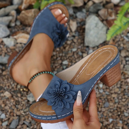 Dusty blue Zylaya women's wedge sandals featuring a layered floral applique, perforated straps, and a wood-grain textured block heel, shown with a foot wearing one sandal and a hand holding the other.