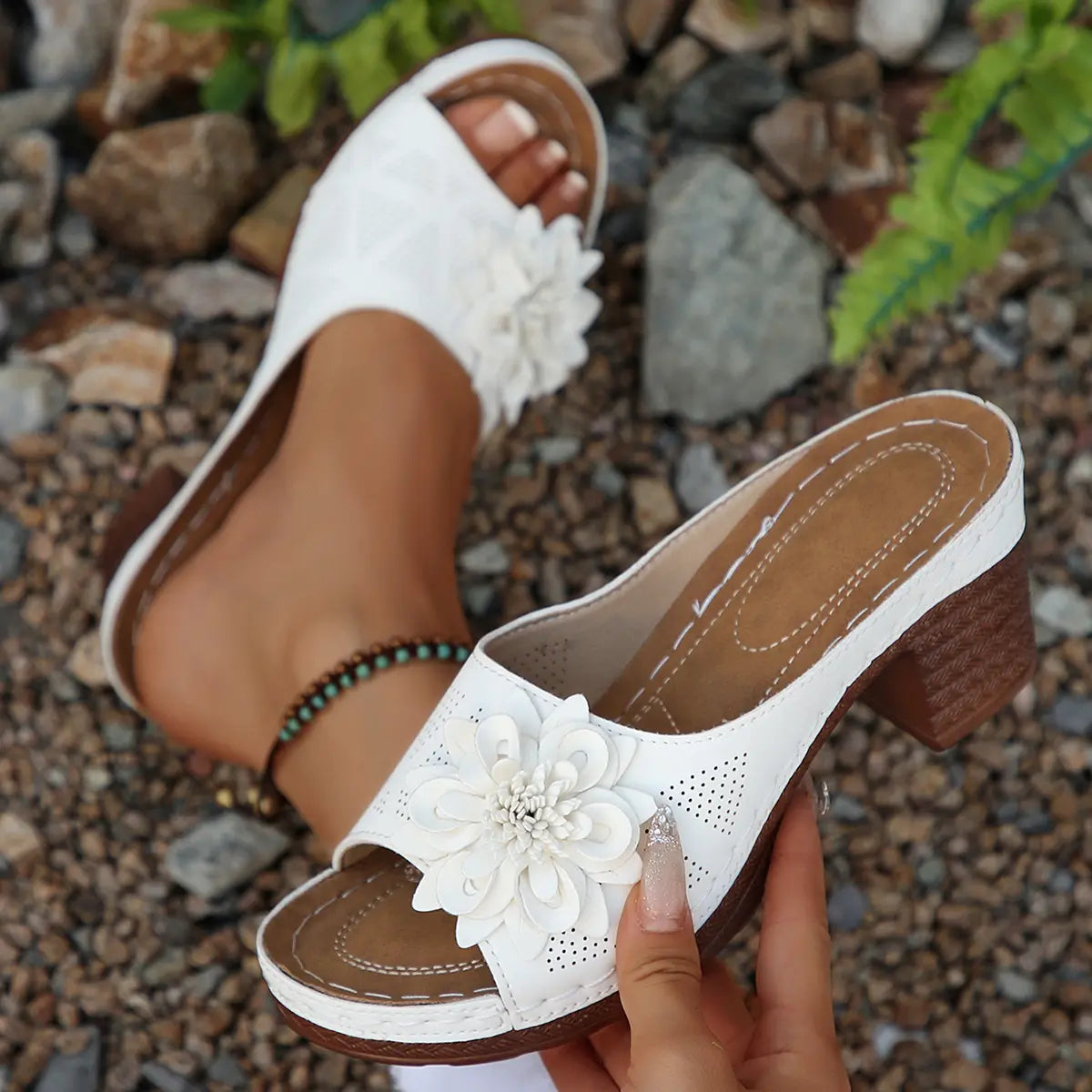 Elegant white Zylaya wedge sandals with a delicate layered floral applique and perforated straps, shown on a gravel path with natural greenery. These slip-on peep-toe sandals feature a comfortable cushioned footbed and a wood-textured block heel.